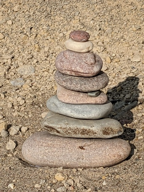 Ein Stapel aus sieben Steinen unterschiedlicher Größe und Farbe, die auf einer kiesigen Oberfläche übereinander balanciert sind.