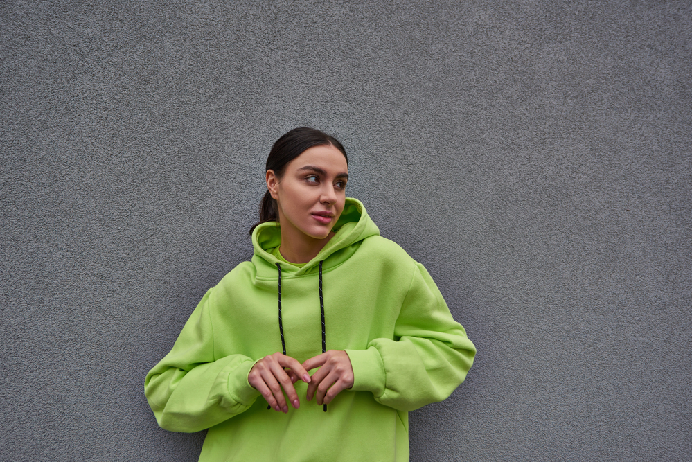 Frau in neongrünem Kapuzenpullover steht vor einer grauen Wand und blickt mit nachdenklichem Gesichtsausdruck weg.