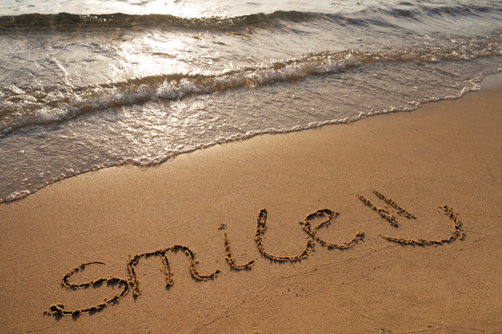 Das Wort „Lächeln!“ ist in den Sand eines Strandes geschrieben, im Hintergrund nähern sich sanfte Wellen.