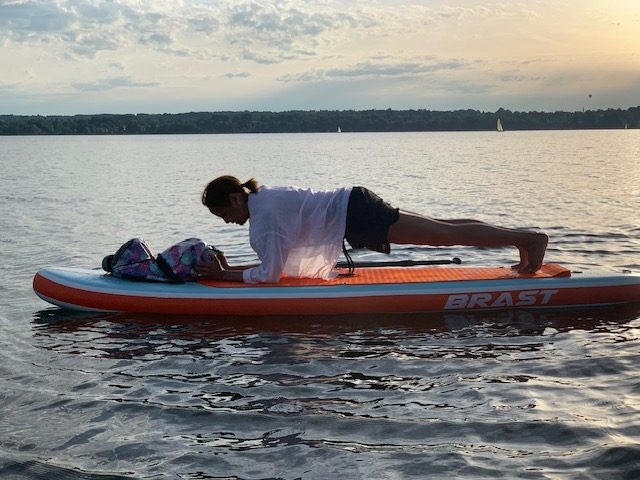 Eine Person, die auf einem orangefarbenen Paddleboard auf einem ruhigen See eine Plank-Übung macht; im Hintergrund sind ein Rucksack und die ferne Uferlinie unter einem bewölkten Himmel zu sehen.