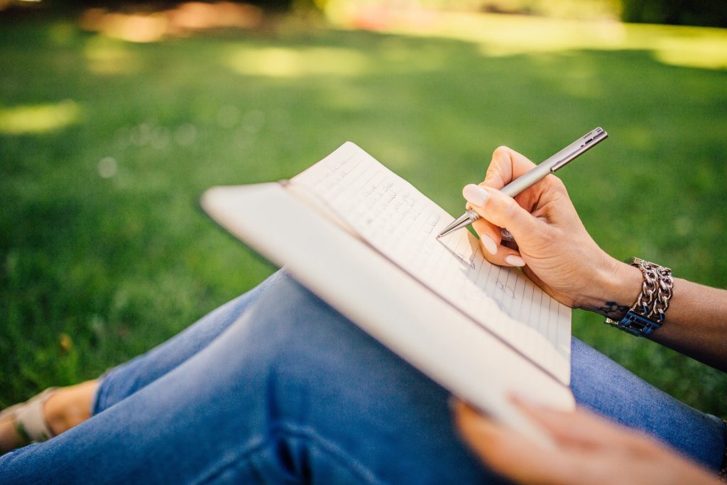 Eine Person, die auf dem Rasen sitzt und mit einem Stift in ein Notizbuch schreibt; sie trägt blaue Jeans und ein Armband und befindet sich draußen in einem sonnigen Park.